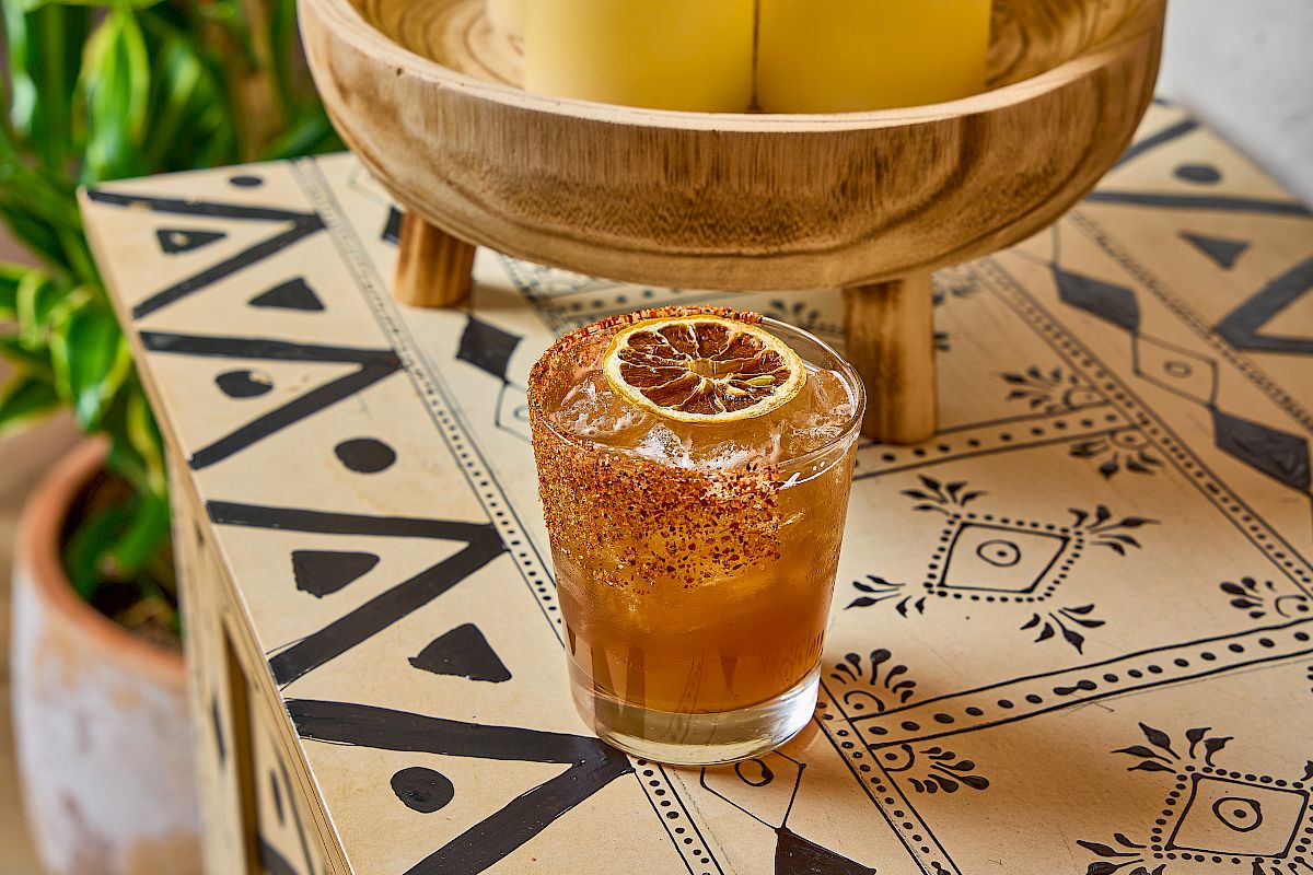 A glass with a drink garnished with a dried citrus slice, on a patterned table next to a wooden bowl with candles.