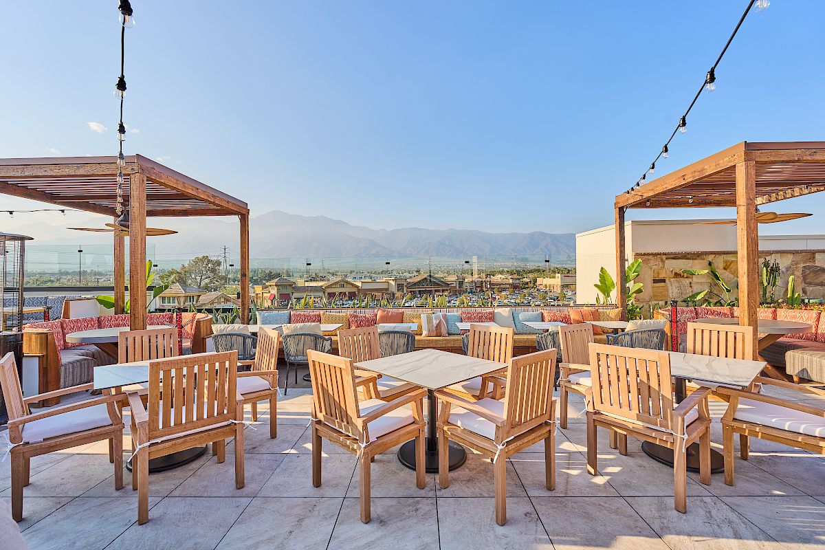 Outdoor seating area with wooden tables and chairs beneath pergolas, overlooking a scenic view with mountains and clear skies in the background.