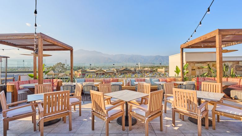 Outdoor seating area with wooden tables and chairs beneath pergolas, overlooking a scenic view with mountains and clear skies in the background.