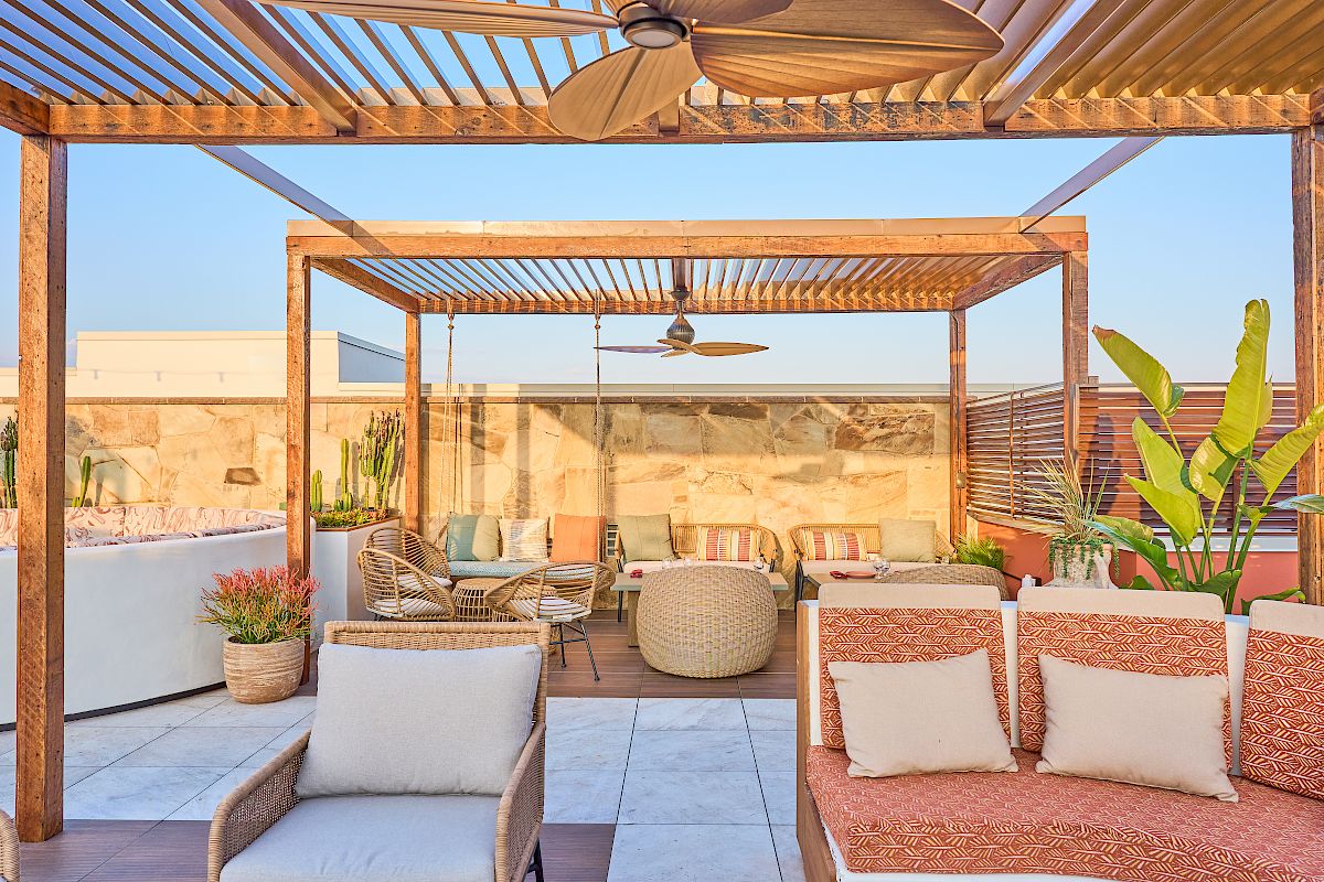 A cozy outdoor patio with pergola, seating, ceiling fan, and plants, set against a clear sky and stone wall backdrop.