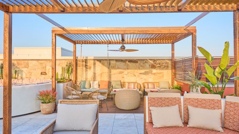 A cozy outdoor patio with pergola, seating, ceiling fan, and plants, set against a clear sky and stone wall backdrop.
