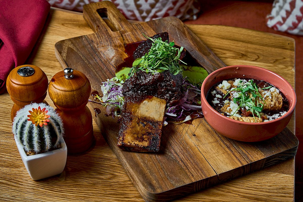 A wooden board with seasoned ribs, greens, and a side bowl of mixed grains. Nearby are salt, pepper shakers, and a small cactus.