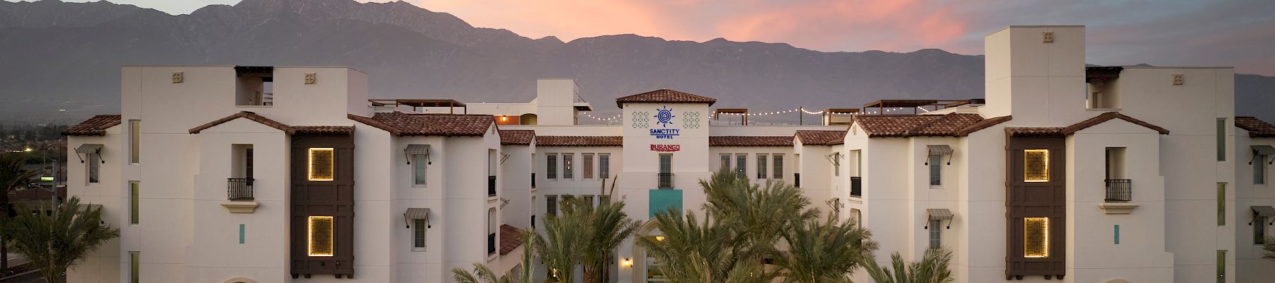 A large building with white walls and a central entrance is surrounded by palm trees, set against a mountain backdrop and colorful sky.