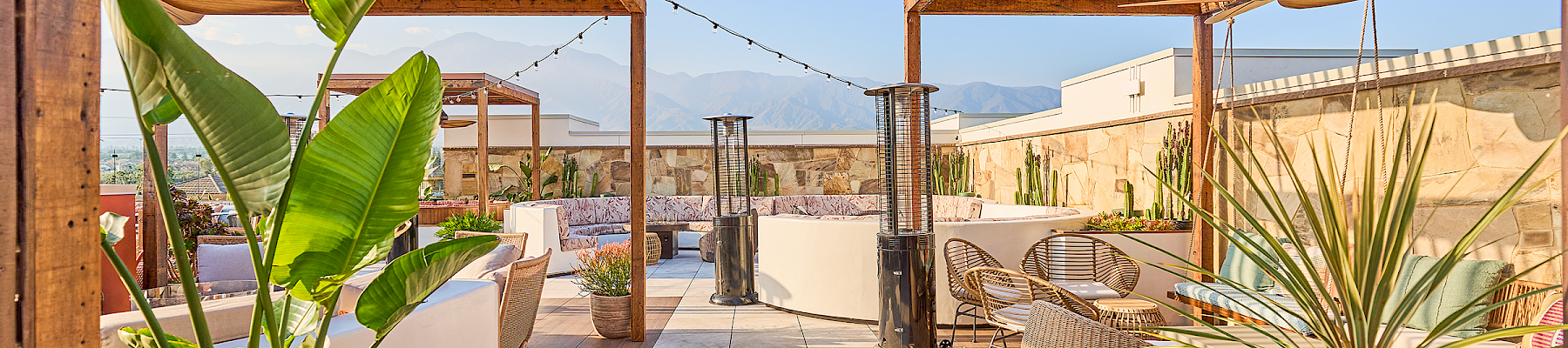 A rooftop patio with pergolas, plants, seating, and heaters offers a cozy outdoor space with mountain views in the background.